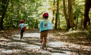Two children wearing a hood while riding a three-wheeled scooter.