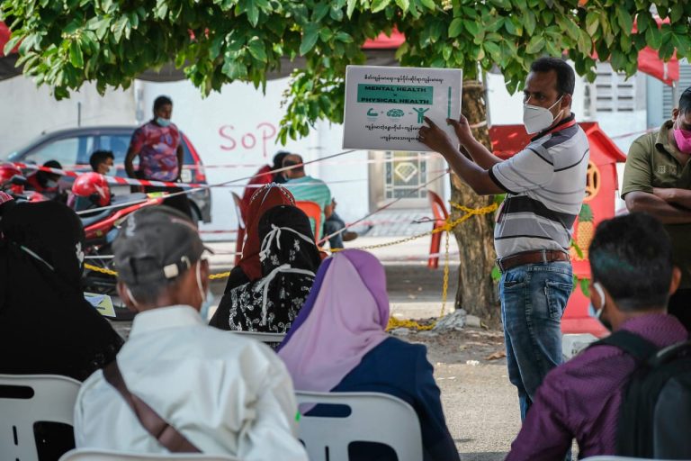 A refugee holding a sign about mental health while speaking to fellow refugees.
