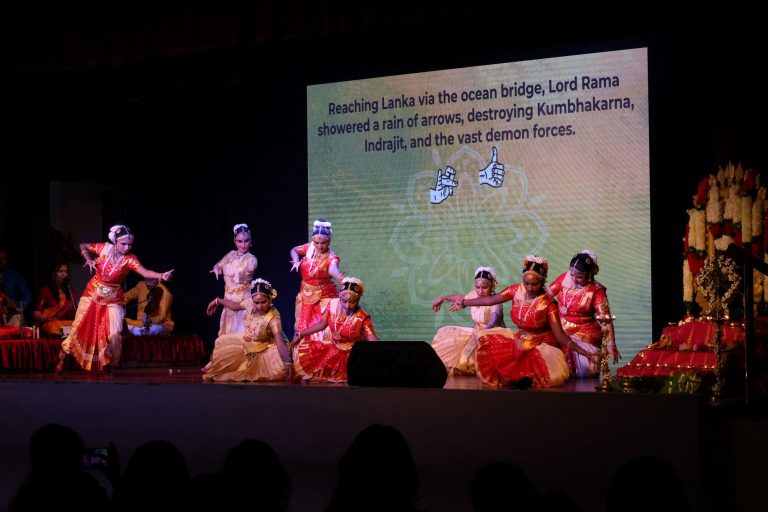 Classical Indian dancers on stage, backed by a large projection screen displaying subtitles and the meaning of hand gestures.